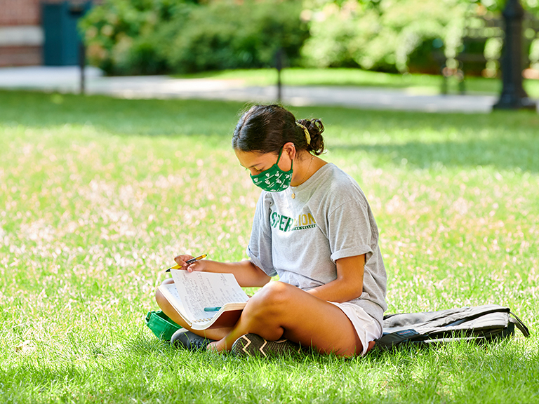 Female student on the quad wearing a mask and reading.